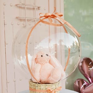 Es un peluche suave dentro de un globo transparente sobre una cesta de presentación decorada con lazos.